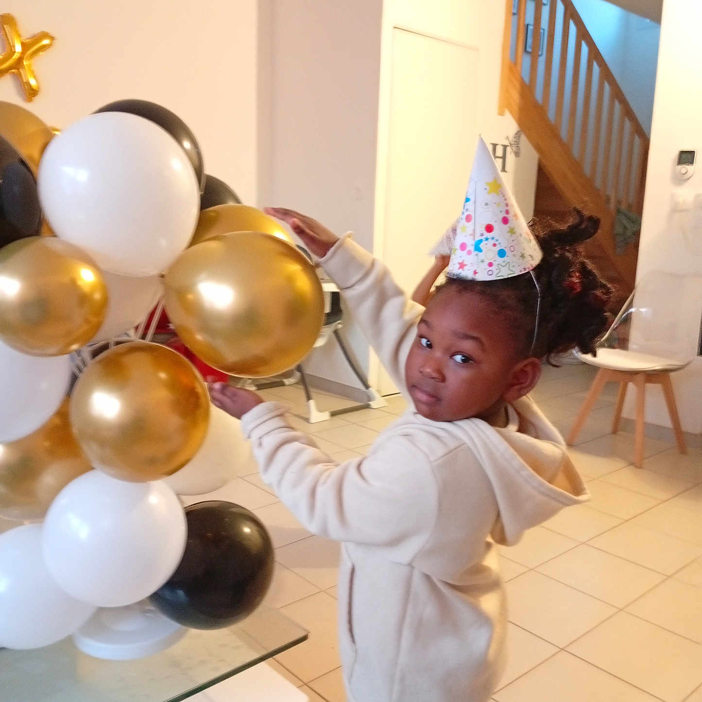 Yloane a rejoint le concours — aidez-le/la à gagner de superbes lots ! balloons, birthday, black_balloons, casual_clothing, celebration, chair, child, curly_hair, glass_table, gold_balloons, home, hoodie, indoor, party_hat, person, smiling, staircase, tile_floor, white_balloons, young_child