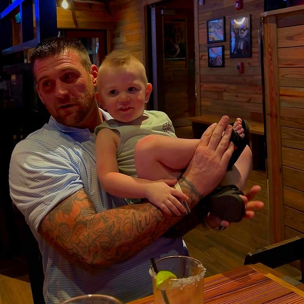 Cash-William joined the competition — help win amazing prizes! arm, beard, casual_clothing, child, drink, facial_expression, family, glass, holding, indoor, lighting, man, mohawk, restaurant, seat, smile, table, tattoos, toddler, wooden_wall