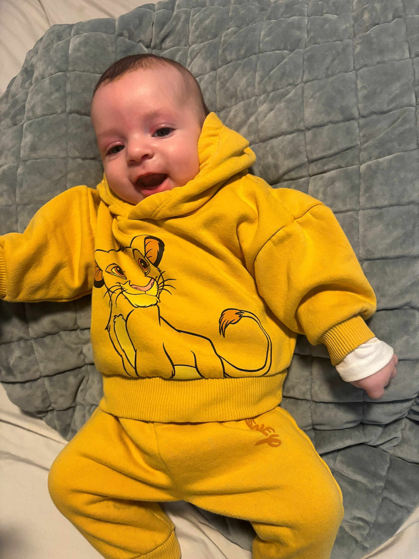 Adrian is registered to the contest to win money with this photo: baby, child, yellow_clothing, hoodie, pants, smile, face, infant, blanket, bed, happy, cute, lying_down, cartoon_character, lion_king, long_sleeve, casual_wear, cozy, indoors, portrait