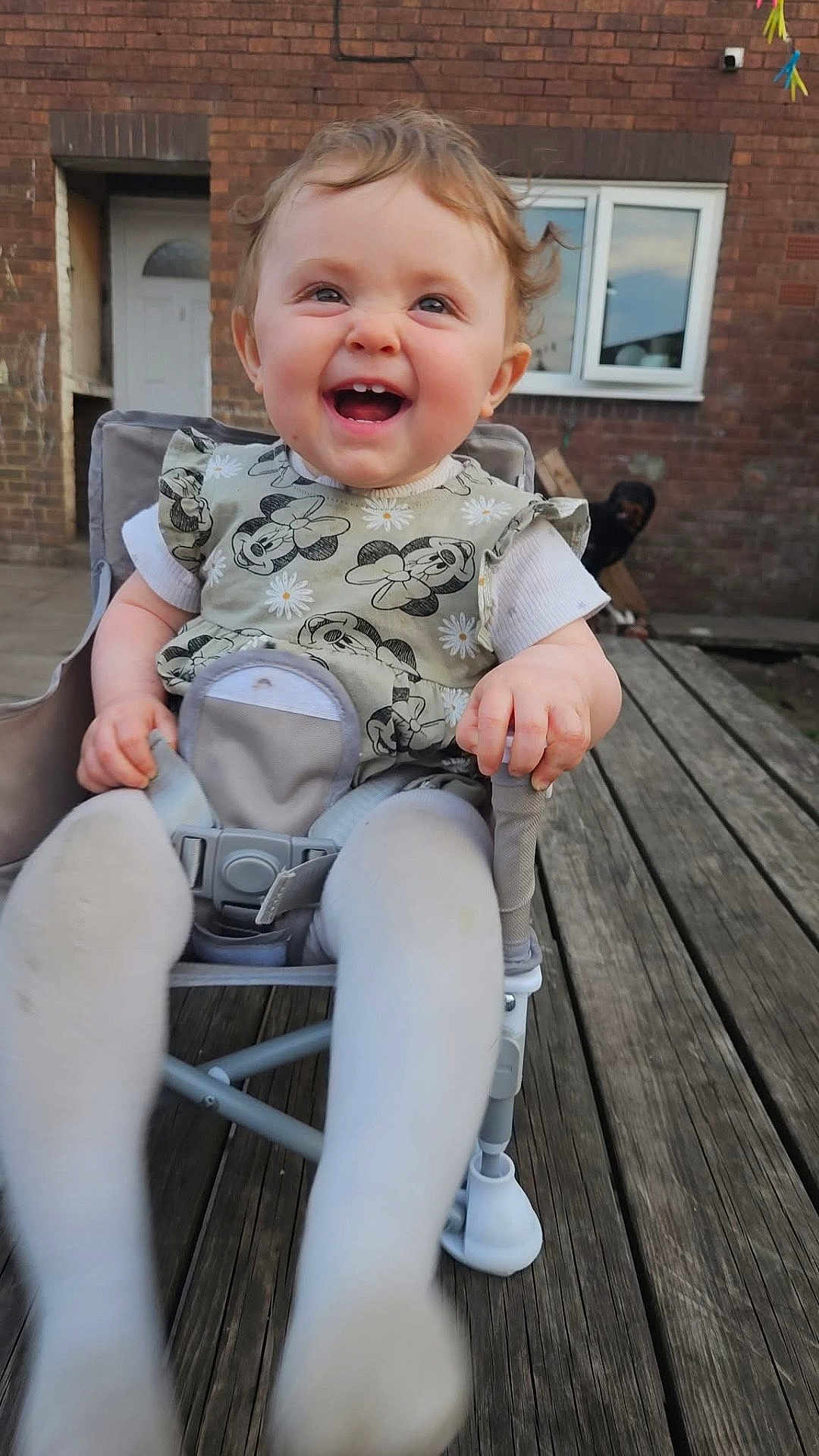 Arabella is registered to the contest to win money with this photo: baby, child, smiling, happy, mickey_mouse, dress, curly_hair, chair, outdoor, wooden_deck, building, window, door, person, tights, feet, hand, seatbelt, playful, young