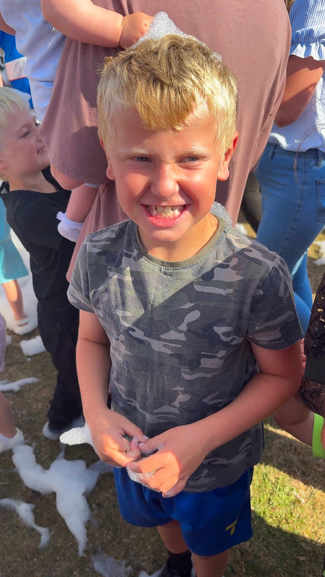 Corey is registered to the contest to win money with this photo: boy, child, blond_hair, camouflage_shirt, blue_shorts, foam, bubbles, outdoor, grass, sunlight, smiling, happy, playful, people, children, summer, fun, group, casual_clothing, daytime