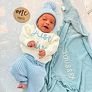 Youssef joined the competition — help win amazing prizes! baby, newborn, infant, one_month_old, portrait, lying_down, smiling, blue_hat, knitwear, sweater, pants, blanket, milestone_card, wooden_sign, nursery, bed, patterned_sheet, personalized_blanket, indoor, safe_sleep_scene