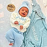 baby, newborn, infant, one_month_old, portrait, lying_down, smiling, blue_hat, knitwear, sweater, pants, blanket, milestone_card, wooden_sign, nursery, bed, patterned_sheet, personalized_blanket, indoor, safe_sleep_scene