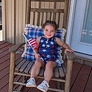 Kira joined the competition — help win amazing prizes! child, rocking_chair, american_flag, porch, patriotic_clothing, plaid_cushion, wooden_furniture, star_decorations, smiling, young_child, outdoor, summer, happy, casual, footwear, braided_hair, decor, wooden_floor, door, home