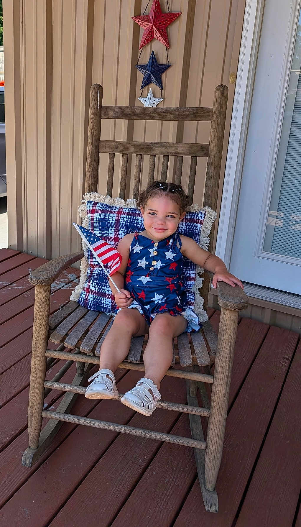 Kira joined the competition — help win amazing prizes! child, rocking_chair, american_flag, porch, patriotic_clothing, plaid_cushion, wooden_furniture, star_decorations, smiling, young_child, outdoor, summer, happy, casual, footwear, braided_hair, decor, wooden_floor, door, home