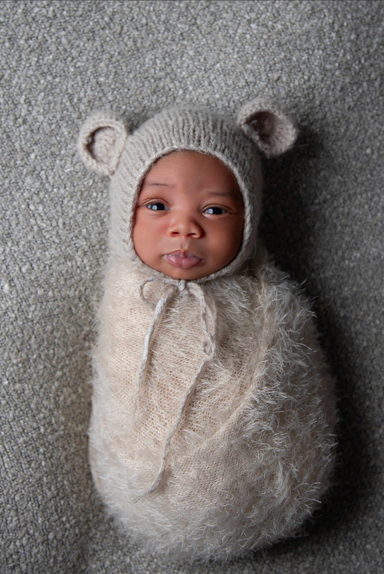 Elijah joined the competition — help win amazing prizes! baby, infant, swaddled, knit_hat, bear_ears, fuzzy_wrap, cute, portrait, looking_at_camera, face, newborn, soft_texture, cozy, lying_down, gray_background, closeup, child, adorable, warm_clothing, indoor