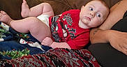 Kelly is registered to the contest to win money with this photo: baby, infant, child, portrait, sitting, lying_down, couch, blanket, red_shirt, diaper, face, eyes, looking_at_camera, indoor, home, adult_arm, tattoo, furniture, cozy, photography