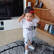 Ezio participe au concours pour gagner de l'argent avec cette photo : child, toddler, cap, white_shirt, shorts, indoor, rug, carpet, living_room, bookshelf, books, framed_photos, wooden_shelf, television, floor, sneakers, pacifier, playful, person, furniture