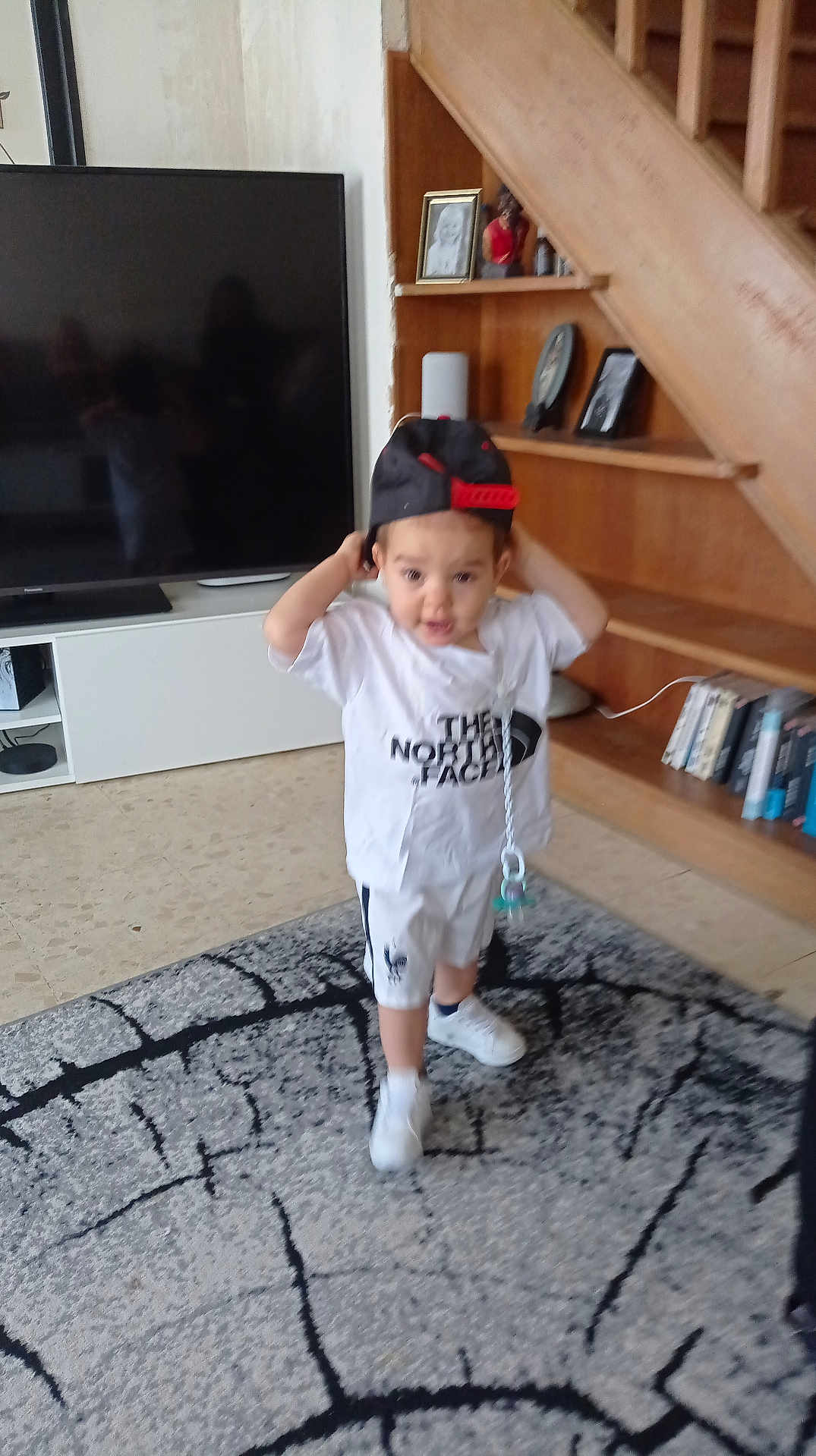 Ezio participe au concours pour gagner de l'argent avec cette photo : child, toddler, cap, white_shirt, shorts, indoor, rug, carpet, living_room, bookshelf, books, framed_photos, wooden_shelf, television, floor, sneakers, pacifier, playful, person, furniture