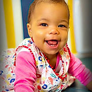 Uhaina Joyaux Mboyo a rejoint le concours — aidez-le/la à gagner de superbes lots ! baby, child, smiling, crawling, colorful_clothing, floral_pattern, pink_sleeve, happy, face, eyes, mouth, teeth, indoor, vibrant_background, striped_background, person, portrait, cute, infant, playful