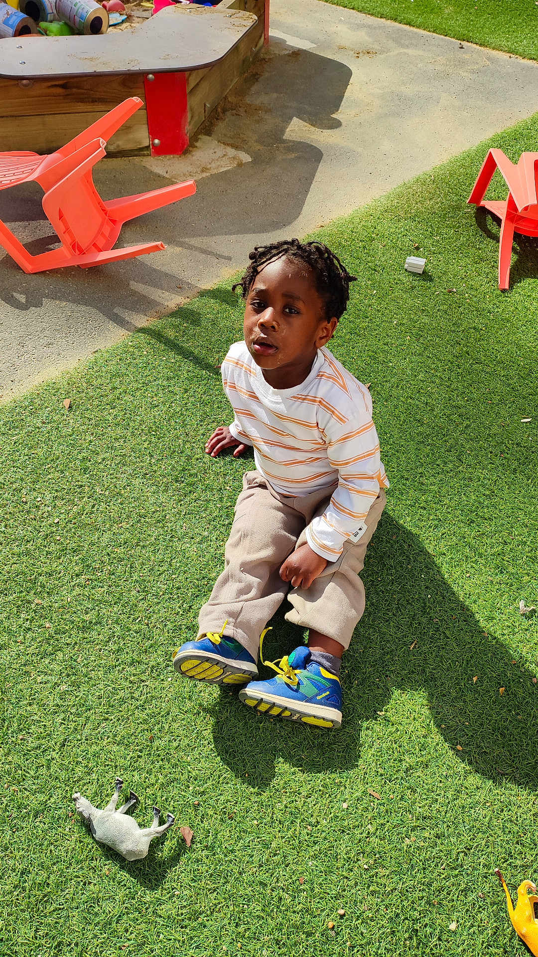 Jezréel-Kendy a rejoint le concours — aidez-le/la à gagner de superbes lots ! child, boy, outdoor, grass, toy, plastic_chair, braided_hair, striped_shirt, shoes, playground, sunlight, shadow, sitting, curious, casual_clothing, beige_pants, blue_shoes, playtime, daylight, greenery