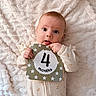 baby, infant, milestone_card, 4_months, blanket, onesie, cute, face, tongue_out, hands, lying_down, soft_texture, patterned_clothing, child, young_baby, indoor, portrait, light_background, playful, cozy