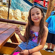 Noursine a rejoint le concours — aidez-le/la à gagner de superbes lots ! child, girl, smiling, outdoor, picnic_table, snack, casual_clothing, shorts, sitting, playground, amusement_park, ride, trees, sunlight, fence, shoes, long_hair, blue_sky, happy, summer