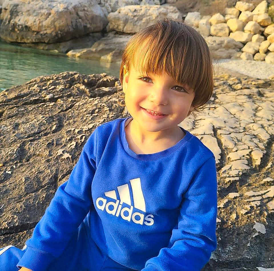 Amir participe au concours pour gagner de l'argent avec cette photo : child, smiling, blue_sweatshirt, adidas_logo, rocks, water, outdoor, sunlight, portrait, happy, casual_clothing, nature, young_child, daylight, scenic, relaxed, face, hair, sitting, playful