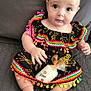 baby, infant, dress, colorful_clothing, pom_poms, candle, wrapped_gift, sitting, gray_background, cute, child, portrait, indoors, jewelry, necklace, footwearless, hand, face, eyes, soft_surface