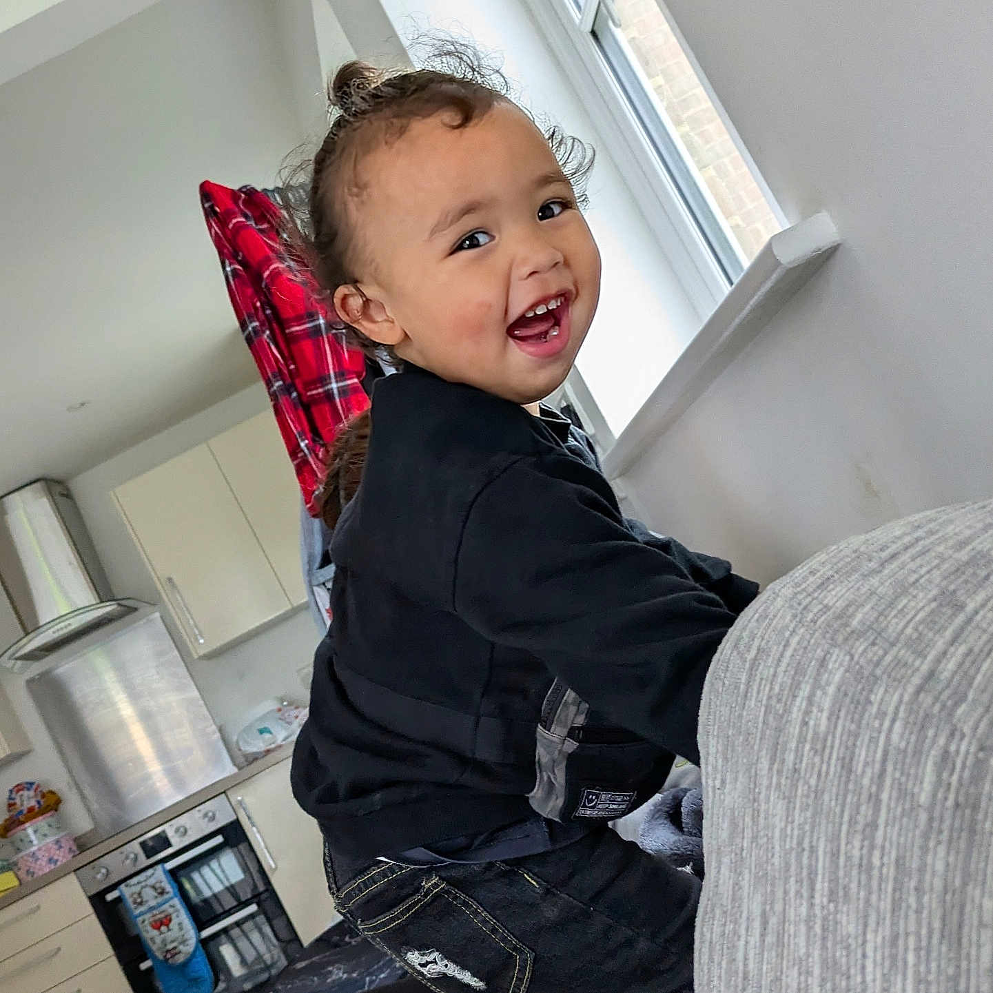 Datu is registered to the contest to win money with this photo: appliances, black_clothing, casual, child, couch, curly_hair, daylight, face, furniture, happy, home, indoor, jeans, kitchen, person, playing, red_plaid, smiling, toddler, window