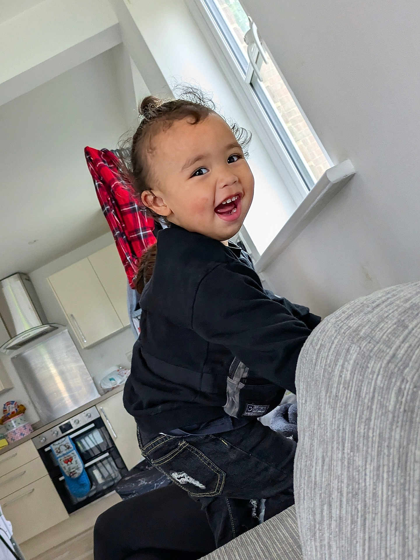 Datu is registered to the contest to win money with this photo: toddler, child, smiling, indoor, kitchen, couch, window, black_clothing, curly_hair, happy, playing, jeans, red_plaid, appliances, home, person, face, casual, furniture, daylight