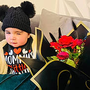 Zayaan joined the competition — help win amazing prizes! toddler, child, winking, hat, pom_poms, jacket, couch, roses, bouquet, flowers, red_roses, black_wrap, gold_trim, indoor, cozy, cute, portrait, expression, warm_clothing, smile