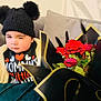 toddler, child, winking, hat, pom_poms, jacket, couch, roses, bouquet, flowers, red_roses, black_wrap, gold_trim, indoor, cozy, cute, portrait, expression, warm_clothing, smile