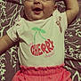 baby, smiling, infant, clothing, pink_shorts, shirt, lying_down, happy, face, arm_raised, bedspread, pattern, cute, child, person, young, indoors, apparel, text_on_clothing, cheerful