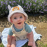 Austin joined the competition — help win amazing prizes! baby, child, hat, striped_hat, lion_patch, outdoor, grass, flowers, purple_flowers, blue_eyes, curious, sitting, cloth, sunlight, nature, cute, infant, smiling, barefoot, young_child