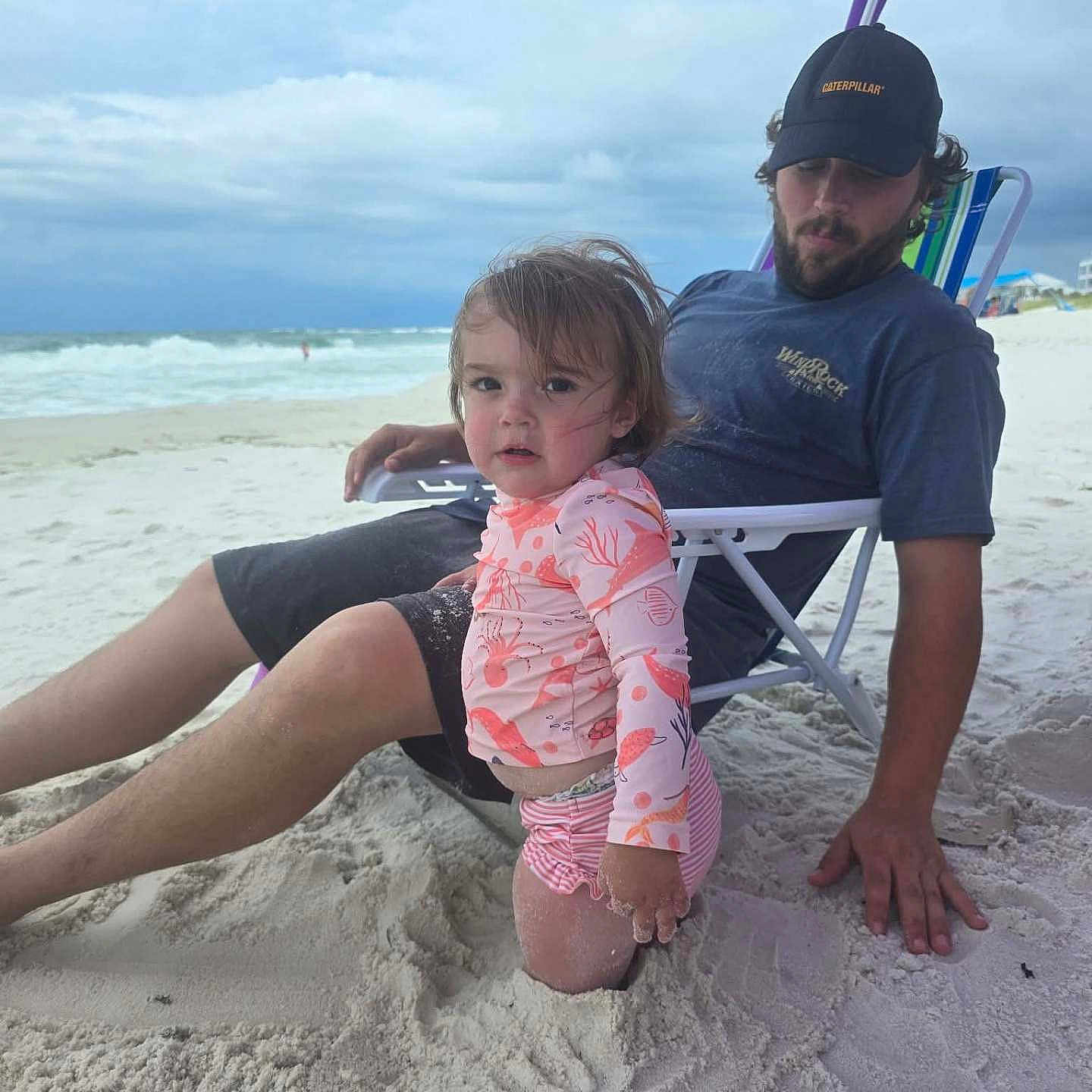 Addeline is registered to the contest to win money with this photo: beach, beach_chair, child, cloudy_sky, hat, man, ocean, outdoor, person, relaxing, sand, shorts, starfish_shaped_sand_mold, summer, swimsuit, toddler, toy, umbrella, vacation, waves