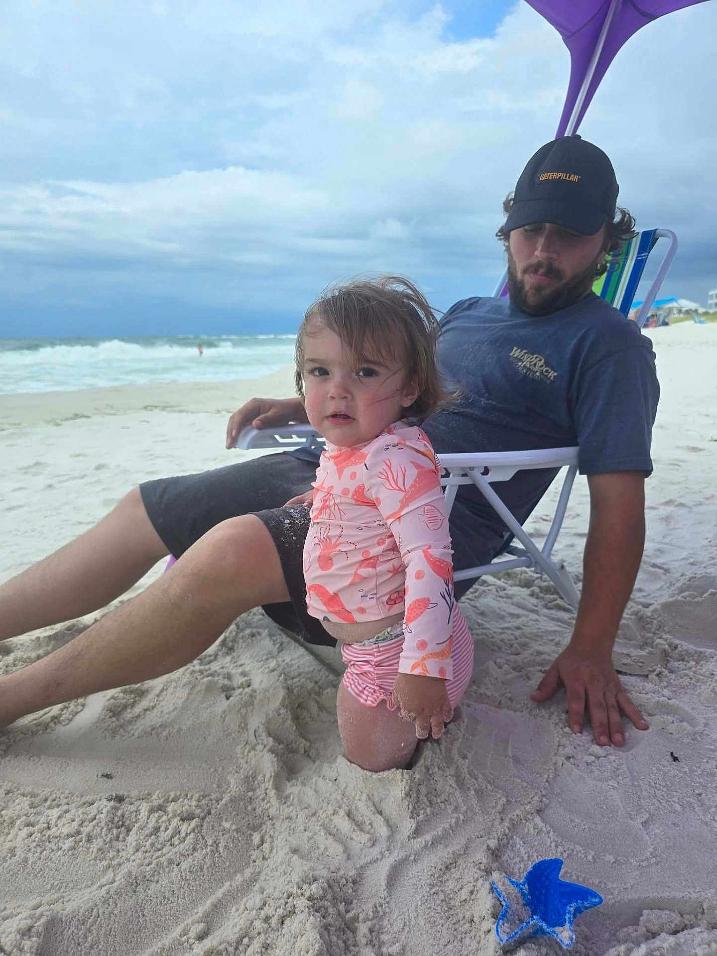 Addeline is registered to the contest to win money with this photo: toddler, child, man, beach, sand, ocean, waves, umbrella, beach_chair, swimsuit, shorts, hat, cloudy_sky, person, relaxing, summer, vacation, outdoor, toy, starfish_shaped_sand_mold