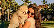Niña is registered to the contest to win money with this photo: woman, dog, pomeranian, sunglasses, outdoor, park, grass, trees, palm_trees, sunlight, smile, happy, pet, summer, casual_clothing, nature, daytime, portrait, cute, animal