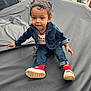 child, toddler, denim_jacket, jeans, red_socks, white_sneakers, curly_hair, bows, smiling, outdoor, black_tarp, sitting, playful, cute, young_child, casual_clothing, person, portrait, daylight, happy