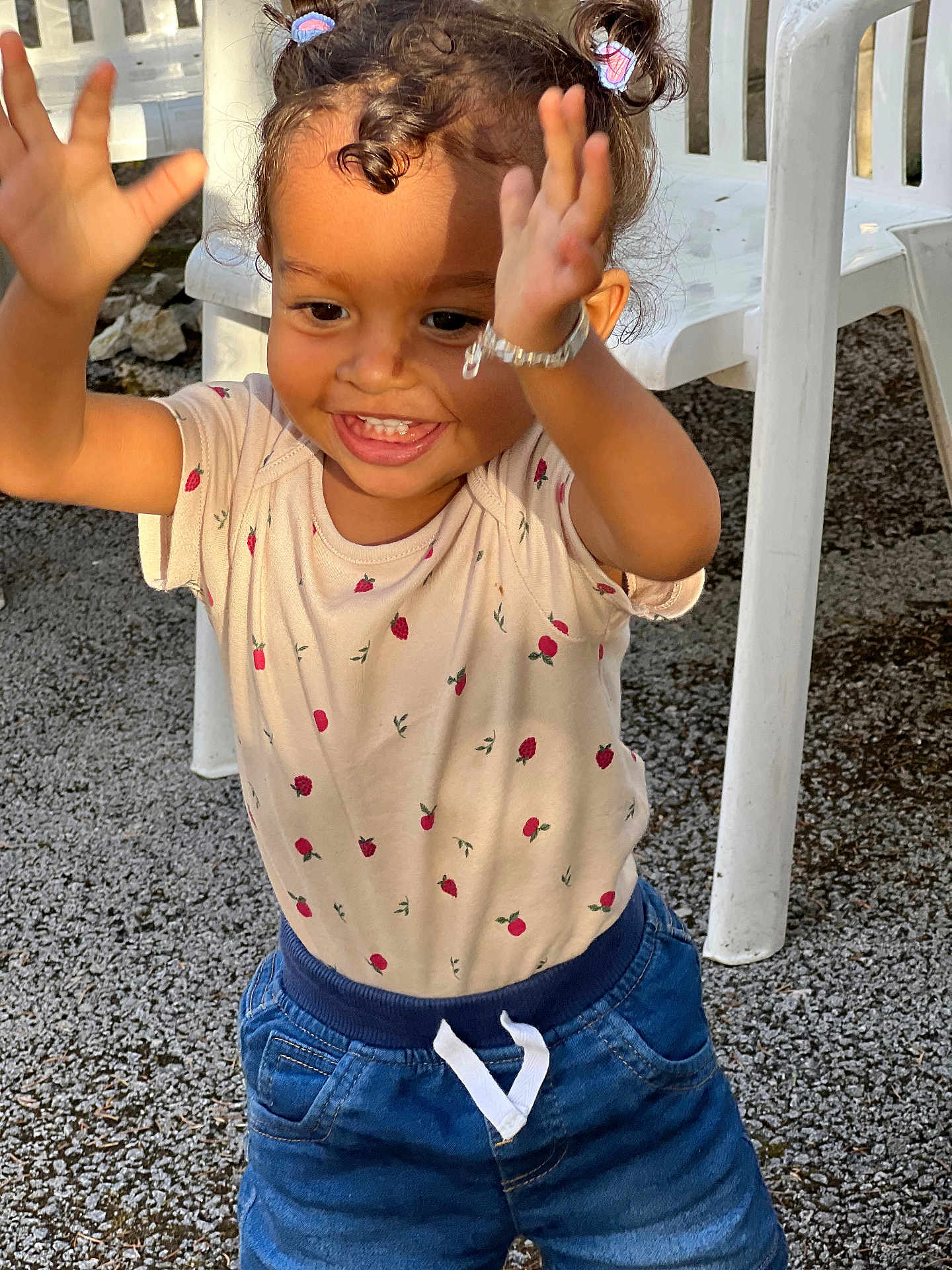 Harena participe au concours pour gagner de l'argent avec cette photo : toddler, child, happy, smiling, curly_hair, pigtails, bracelet, shorts, shirt, strawberry_pattern, outdoor, plastic_chair, gravel, sunlight, playful, cute, portrait, person, clothing, joy