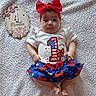 baby, infant, child, headband, red_bow, white_shirt, blue_skirt, 4_months_sign, milestone, blanket, patterned_clothing, patriotic, stars, fireworks_print, cute, lying_down, decor, celebration, portrait, indoors