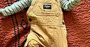 Teddy is registered to the contest to win money with this photo: baby, child, overalls, yellow_clothing, striped_shirt, quilt, textured_bedspread, foot, hand, face, infant, lying_down, patterned_background, cute, young_child, indoor, person, portrait, small_child, relaxed
