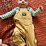 baby, child, overalls, yellow_clothing, striped_shirt, quilt, textured_bedspread, foot, hand, face, infant, lying_down, patterned_background, cute, young_child, indoor, person, portrait, small_child, relaxed