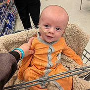 Adam is registered to the contest to win money with this photo: baby, infant, shopping_cart, orange_onesie, striped_clothing, fleece_blanket, nasal_tube, hospital_care, supermarket, aisle, hand, person, floor, clothing, cute, child, face, smiling, indoor, support