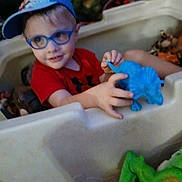 Leviticus is registered to the contest to win money with this photo: child, boy, glasses, cap, toy, dinosaur_toy, playpen, indoor, smiling, red_shirt, blue, plastic, playtime, cute, young_child, person, fun, happy, hands, casual