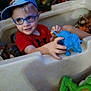 child, boy, glasses, cap, toy, dinosaur_toy, playpen, indoor, smiling, red_shirt, blue, plastic, playtime, cute, young_child, person, fun, happy, hands, casual