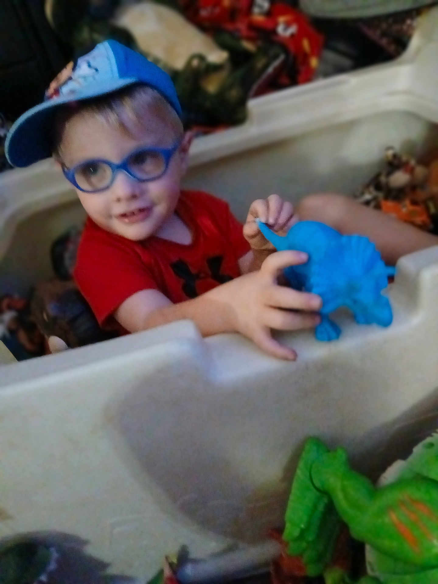 Leviticus is registered to the contest to win money with this photo: child, boy, glasses, cap, toy, dinosaur_toy, playpen, indoor, smiling, red_shirt, blue, plastic, playtime, cute, young_child, person, fun, happy, hands, casual