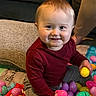 toddler, child, smiling, easter_eggs, colorful, plastic_eggs, indoor, cushion, couch, red_shirt, gray_pants, sitting, playful, happy, baby, person, face, toy, home, floor_mat