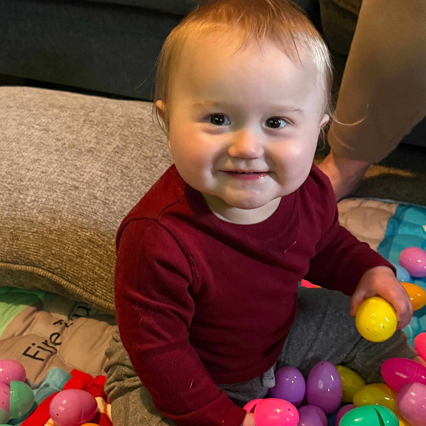 Drew joined the competition — help win amazing prizes! toddler, child, smiling, easter_eggs, colorful, plastic_eggs, indoor, cushion, couch, red_shirt, gray_pants, sitting, playful, happy, baby, person, face, toy, home, floor_mat