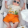 baby, sunglasses, stroller, orange_shorts, animal_print, toy, blanket, pacifier, infant, cute, relaxing, child, clothing, face, headwear, indoors, comfort, person, smiling, casual