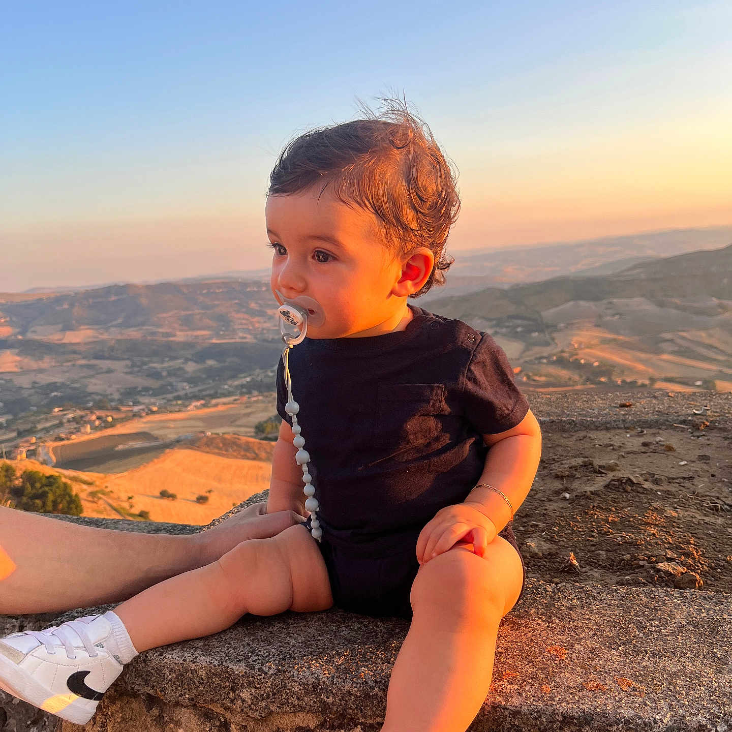 Gabriel participe au concours pour gagner de l'argent avec cette photo : baby, black_clothing, bracelet, child, curly_hair, cute, footwear, hill, landscape, nature, outdoor, pacifier, person, scenery, sitting, sky, sneakers, stone, sunset, toddler