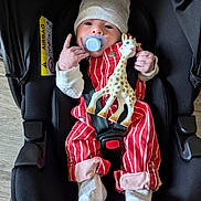 Leo participe au concours pour gagner de l'argent avec cette photo : baby, infant, car_seat, pacifier, hat, toy, giraffe_toy, striped_clothing, white_socks, cute, child, indoors, seatbelt, hand_gesture, newborn, smiling, cuddly, soft_toy, red_clothing, black_car_seat