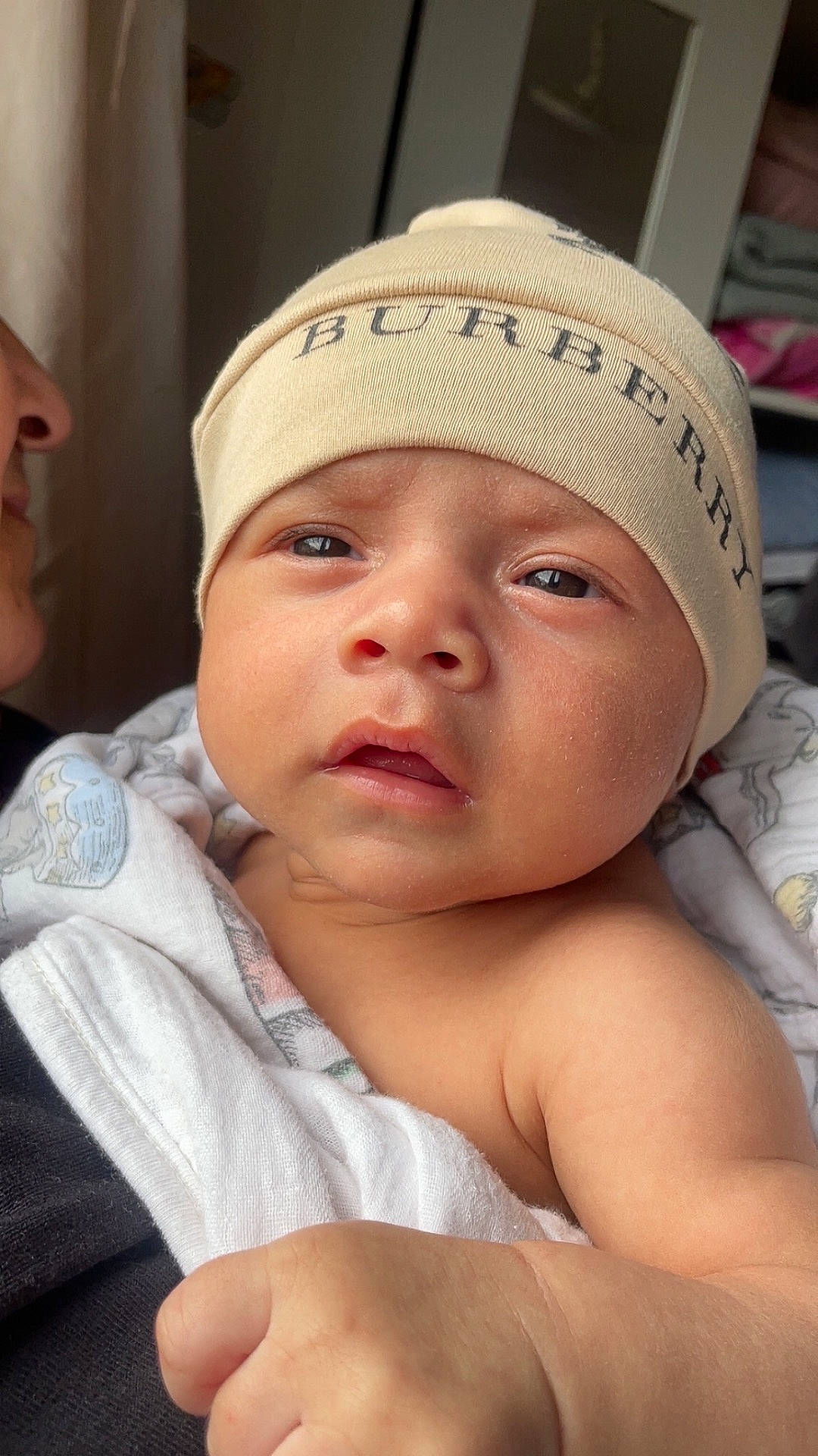 Damian is registered to the contest to win money with this photo: adult, baby, blanket, burberry, close_up, clothing, curious, cute, expression, face, hat, holding, indoors, infant, newborn, person, portrait, skin, soft_light, wrapped