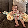 baby, smiling, couch, pillow, infant, clothing, floral_pattern, support_pillow, happy, indoor, person, young_child, sitting, barefoot, cute, comfortable, home, relaxed, colorful_outfit, infant_toddler