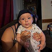 Arizona is registered to the contest to win money with this photo: baby, child, infant, cap, drool, person, hand, indoor, couch, curtain, wall_clock, clothing, face, holding, adult, living_room, furniture, portrait, cute, home