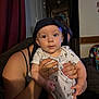 baby, child, infant, cap, drool, person, hand, indoor, couch, curtain, wall_clock, clothing, face, holding, adult, living_room, furniture, portrait, cute, home