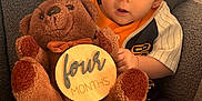 Declan is registered to the contest to win money with this photo: baby, infant, teddy_bear, plush_toy, wooden_sign, four_months, chair, gray_texture, cute_outfit, child, sitting, barefoot, portrait, indoor, curious_expression, soft_toy, cozy, young_baby, person, close_up