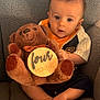 baby, infant, teddy_bear, plush_toy, wooden_sign, four_months, chair, gray_texture, cute_outfit, child, sitting, barefoot, portrait, indoor, curious_expression, soft_toy, cozy, young_baby, person, close_up