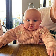 Isla is registered to the contest to win money with this photo: baby, infant, child, pink_sweater, ribbon_bows, wooden_table, table, drool, chubby_cheeks, big_eyes, indoor, window, glass_jar, adult_partial, hand, person, portrait, casual_clothing, restaurant, daylight