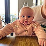 baby, infant, child, pink_sweater, ribbon_bows, wooden_table, table, drool, chubby_cheeks, big_eyes, indoor, window, glass_jar, adult_partial, hand, person, portrait, casual_clothing, restaurant, daylight